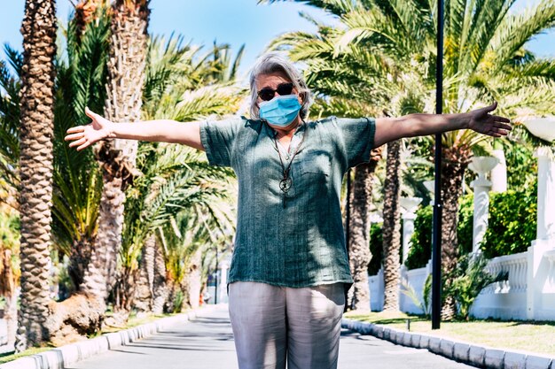 Foto donna adulta anziana che indossa una maschera protettiva a braccia aperte nella strada deserta - blocco a causa del coronavirus covid-19. nessuna gente intorno - palme