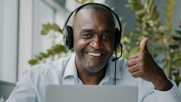 Senior african employee office wear headphones business call on laptop online does thumb up approval