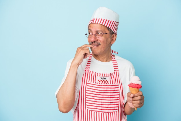 Senior american ice cream man holding an ice cream isolated on blue background relaxed thinking about something looking at a copy space.