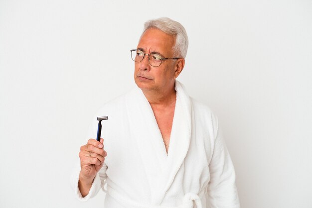 Senior american man wearing bathrobe holding razor blade isolated on white background confused, feels doubtful and unsure.