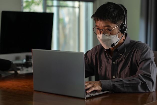 Senior asian businessman in headset and eyeglasses wearing face mask during online video conference sitting at home work from home studying online concept