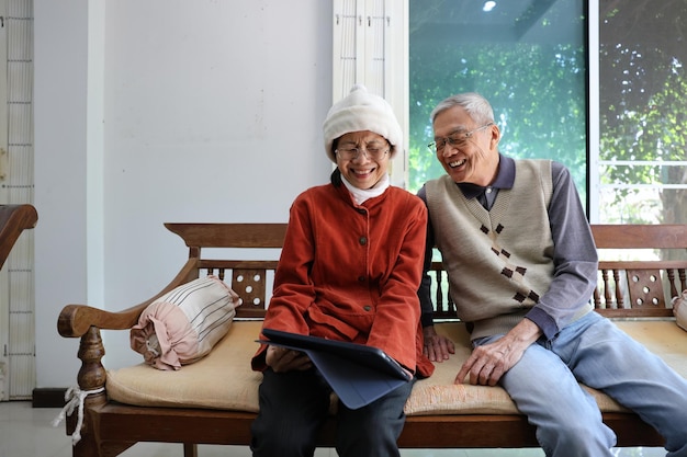 senior asian couple are laughing and sitting on the sofa while watching funny stand up comedy show from streaming digital tablet during winter time for mental health and happiness in marriage