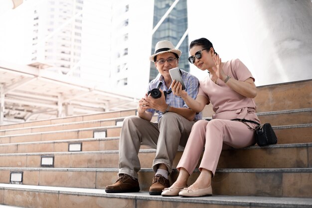 Senior Asian couples sit on the stairs while traveling and have fun video calling.