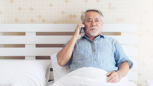 写真 アジアのシニア男性退職した老人は寝室のベッドで携帯電話に電話をかける彼は深刻で心配なライフスタイルをストレスに感じていた自宅のコンセプト