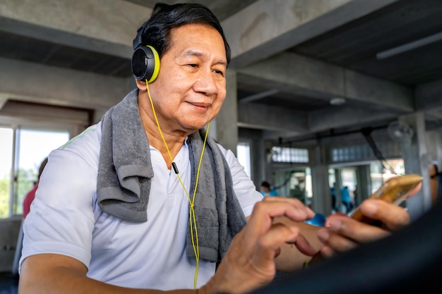 Senior Asian man in sportswear listen to music and training biking cardio at fitness gym.