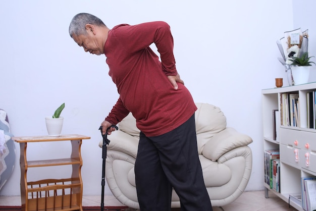 自宅で背中と腰の痛みに苦しんでいる杖を持つ年配のアジア人男性