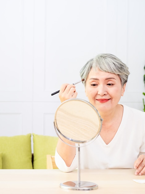 Старшая азиатская женщина наносит основу на щеку с помощью кисти для макияжа, сидя в одиночестве перед зеркалом.