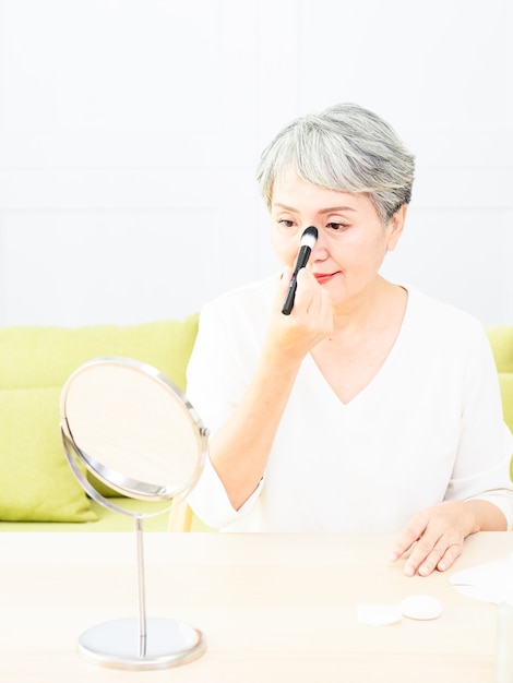 Senior asian woman applying foundation to her cheek with a makeup brush while sitting alone in front of a mirror.
