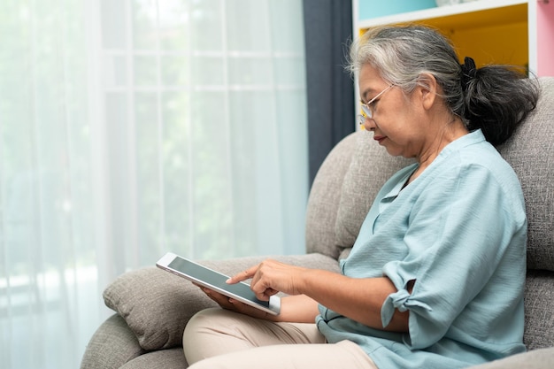 Пожилая азиатка в очках отдыхает дома на диване и использует планшет для чтения новостей