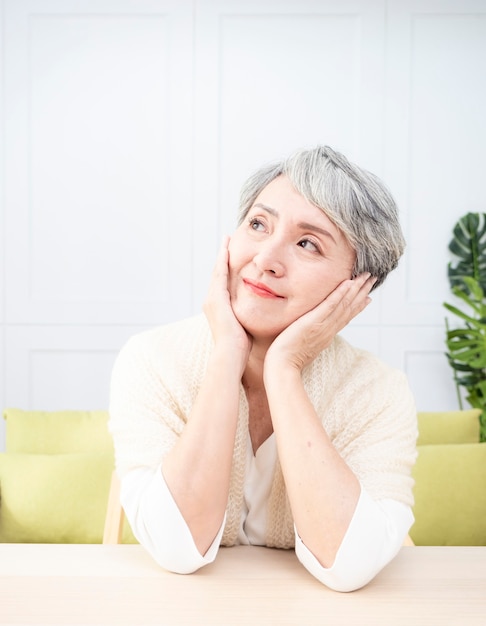 Senior asian woman with short grey hairstyle putting chin on hand and sitting at wood table.