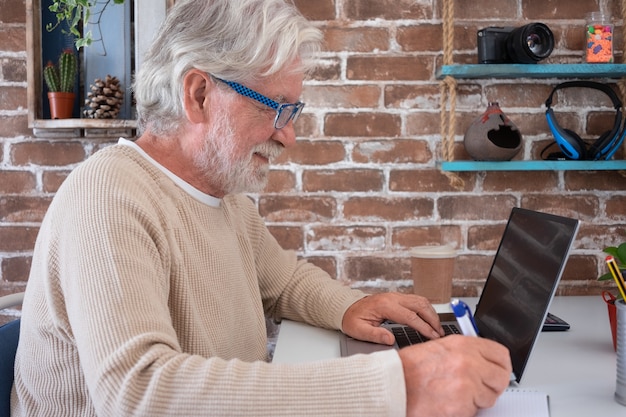 Senior attractive man using laptop computer at home smiling. Brick's wall on background