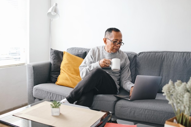 Senior Aziatische man die op de bank zit en een laptop gebruikt terwijl hij thuis een kop koffie vasthoudt