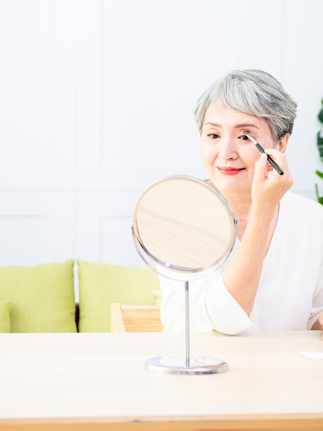 Senior aziatische vrouw die foundation op haar wang aanbrengt met een make-upborstel terwijl ze alleen voor een spiegel zit.