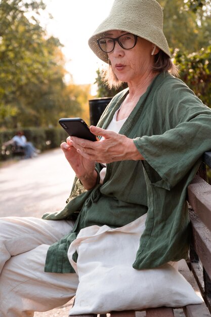 Senior aziatische vrouw in hoed en bril en linnen slijtage spreken tijdens videogesprek in stadspark