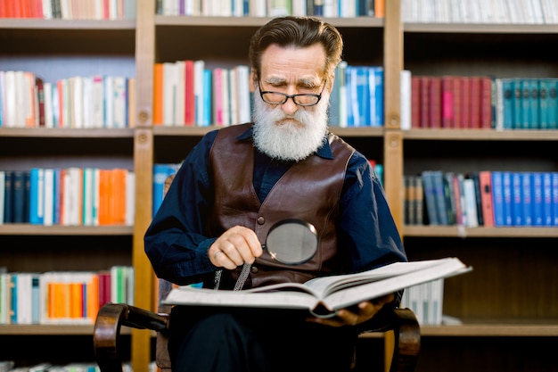 Senior bearded man in glasses, sitting and reading an old book in the library, holding magnifying glass. Knowledge, learning and education concept