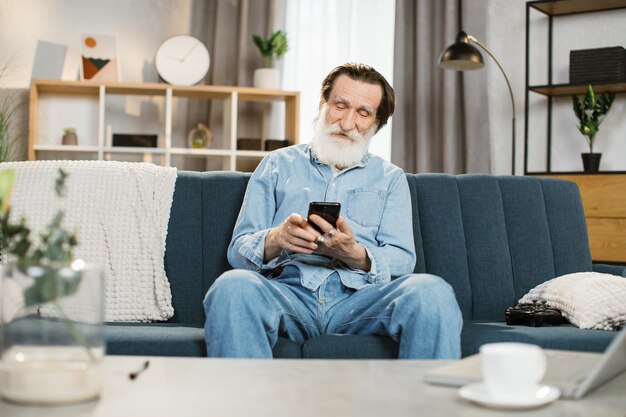 Senior bearded man sitting on the couch at cozy living room\
indoors and using smartphone