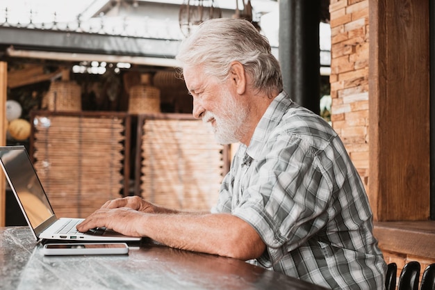 Senior bearded man using laptop sitting outdoor on a wooden table Handsome elderly male browsing internet on laptop