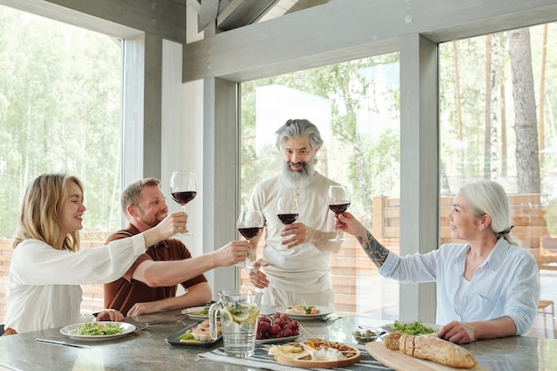 Senior bearded man with glass of red wine drinking toast with family members during dinner