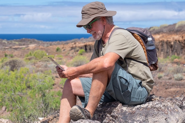 Uomo barbuto anziano con cappello e zaino seduto sulle rocce che riposano dopo il trekking nell'orizzonte di campagna sul mare sullo sfondo