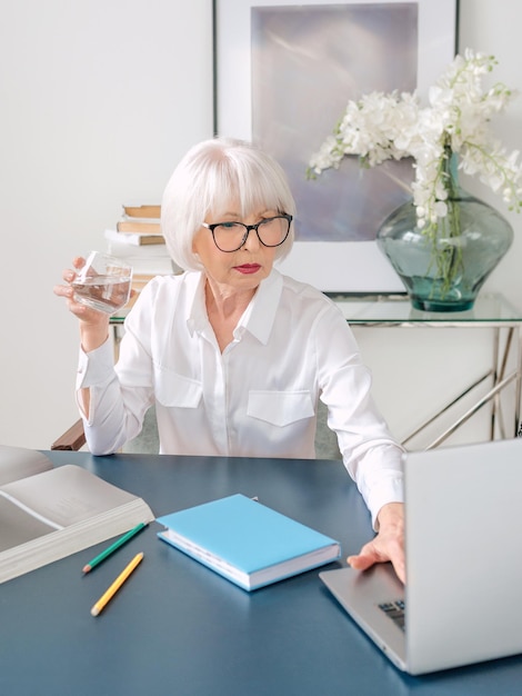 senior beautiful gray hair woman in white blouse drinking water during work in office