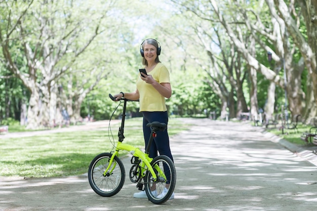 公園を散歩する年配の美しい女性が自転車に乗るヘッドフォンで音楽を聴く電話を持っている彼は自転車を手に持って立ってカメラの笑顔を見る