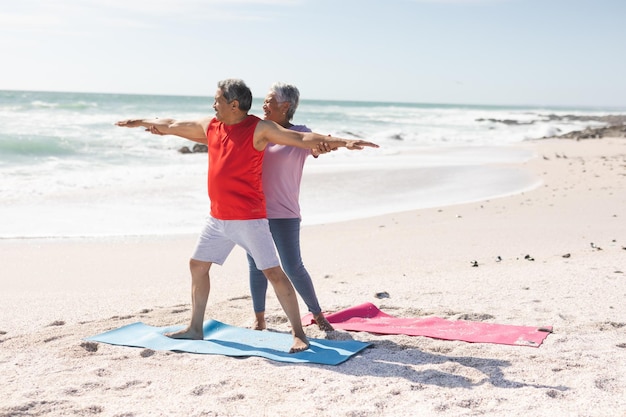 Senior biracial woman helping man with warrior pose yoga posture practicing at beach against sky