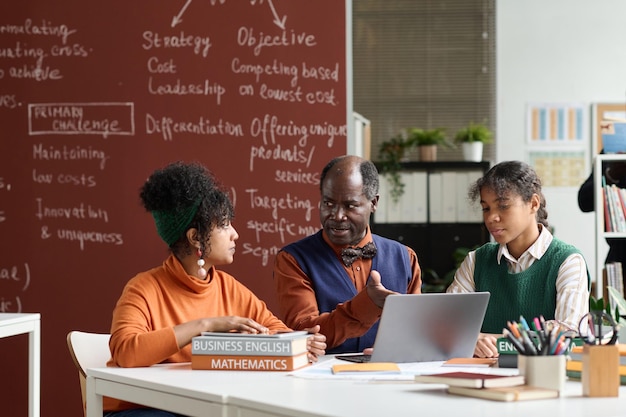 Photo senior black teacher talking to students in class and pointing to laptop