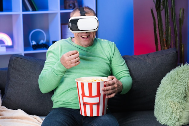 Senior blanke man met een doos popcorn in zijn hand kijken naar video met behulp van Virtual Reality Headset zittend op de bank eten popcorn in de kamer.