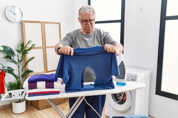Senior blanke man strijken met verbrand ijzeren shirt in wasruimte depressief en zorgen voor nood, huilen, boos en bang. trieste uitdrukking.