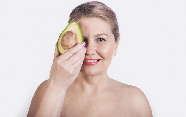 Senior blonde vrouw met naakte schouders die haar gezicht bedekken met een avocadoglimlach