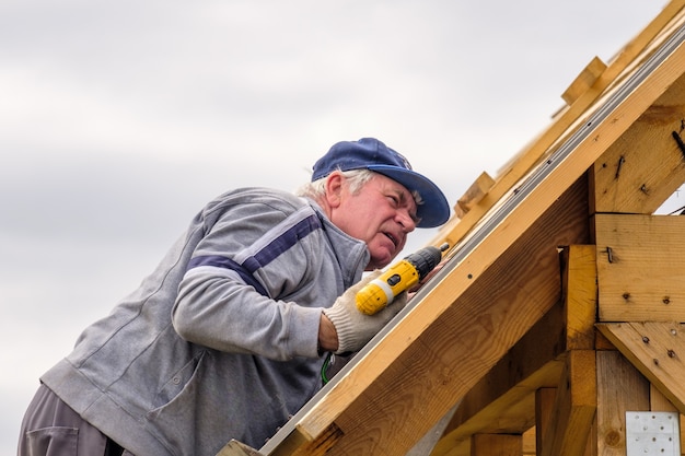 Senior builder man with a screwdriver screwing a roofing sheet\
to the roof