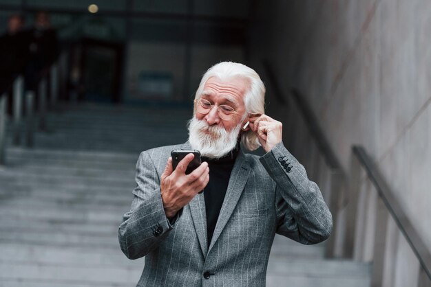 Senior businessman in formal clothes with grey hair and beard is outdoors trying wireless headphones with phone