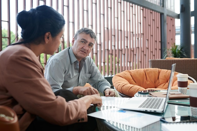 Senior businessman listening to female colleague explaining details in report she prepared for meeting