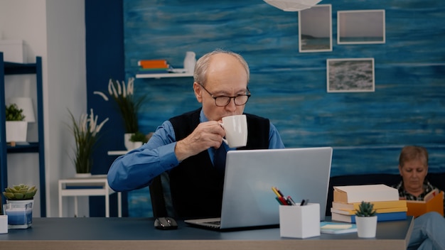 Senior businessman reading reports sitting in front of laptop working from home drinking coffee. Retired man using modern technology analysing typing searching while wife sitting on couch reading a bo