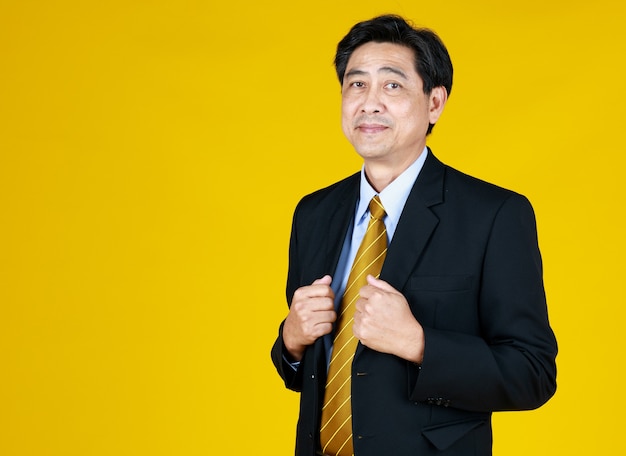 Senior businessman stand, touching black suit. smiling look camera, studio shoot with yellow background. face feel relaxing, happy. concept for ceo, big boss