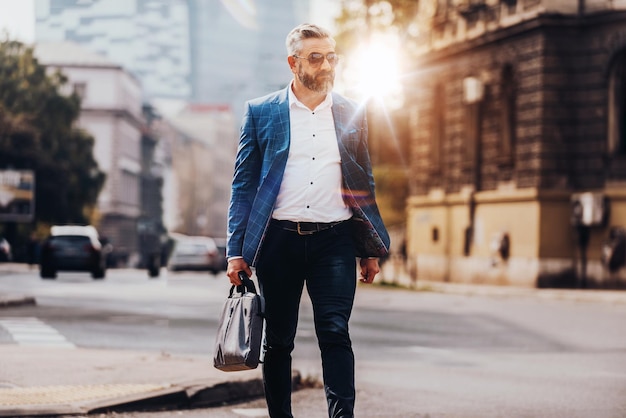 A senior businessman in a suit and sunglasses walking around the city while carrying a bag