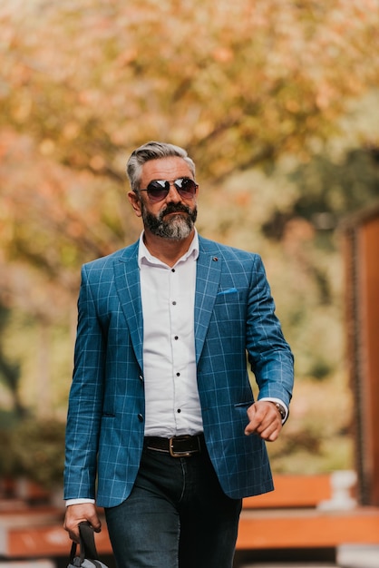 A senior businessman in a suit and sunglasses walking around the city while carrying a bag.