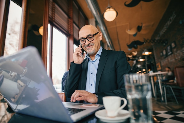 Uomo d'affari senior che utilizza smart phone e computer portatile mentre sedendosi nel caffè