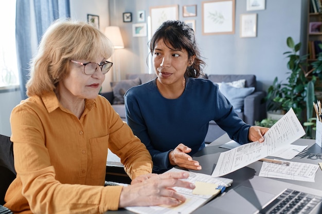 Senior businesswoman in eyeglasses working on laptop with documents at the table together with assistant, they working at home