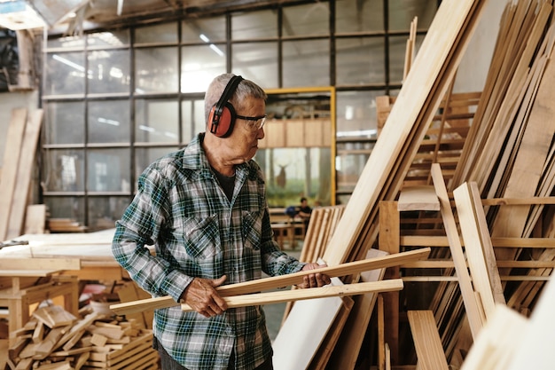 Senior carpenter in earmuffs and plaid shirt carrying wooden planks to workshop storage