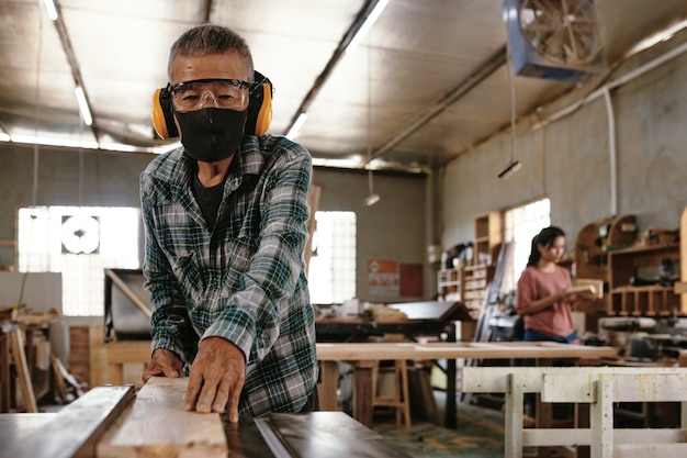 ワークベンチで長い木の板を切る保護フェイスマスクとイヤーマフのシニア大工