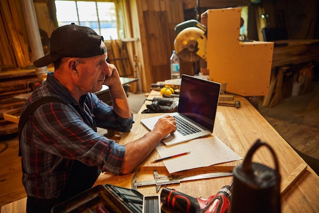 Senior carpenter works on the computer and talks phone with client in the workshop