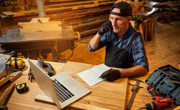 Senior carpenter works on the computer and talks phone with client in the workshop