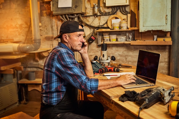 Senior carpenter works on the computer and talks phone with\
client in the workshop