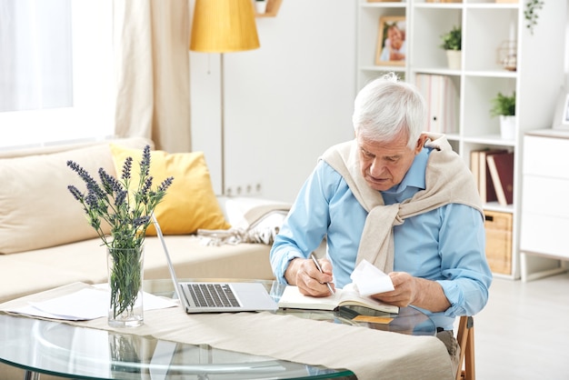 ノートパソコンの前のテーブルで自宅で作業しながらノートにメモをとる白い髪のシニアカジュアルな男
