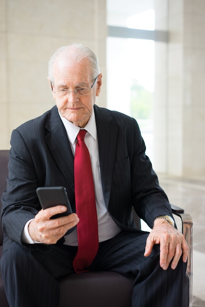 senior Caucasian businessman wearing glasses sitting in armchair and texting 