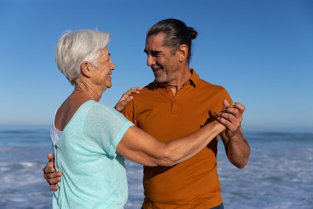 Senior Caucasian couple enjoying time at the beach on a sunny day, dancing together barefoot with sea in the background