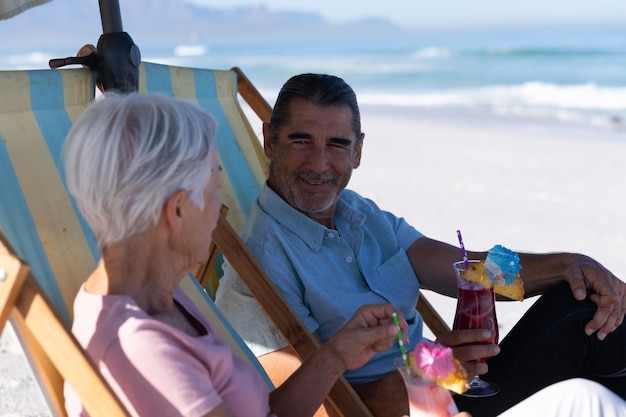 Photo senior caucasian couple enjoying time at the beach on a sunny day, sitting on deck chairs, drinking drinks with sand in the background