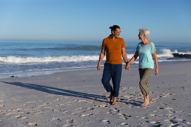 Senior Caucasian couple enjoying time at the beach on a sunny day, walking and holding hands with sea in the background