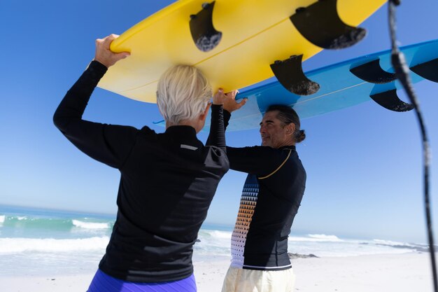 Photo senior caucasian couple enjoying time at the beach, walking towards the sea and holding surfboards above their heads